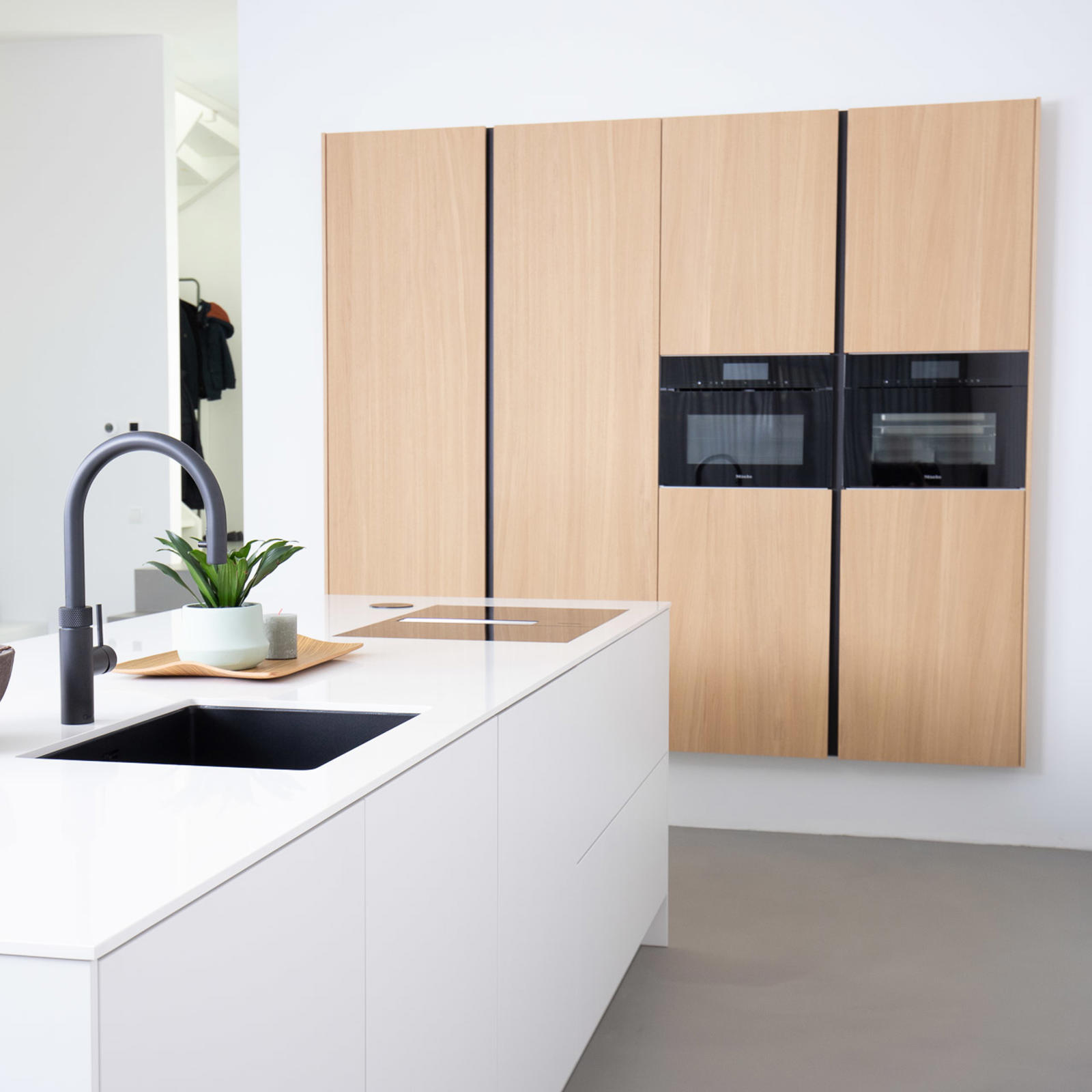 Strakke greeploze keuken in Eijsden: wit eiland met zwarte kraan en zwevende houtlook kastenwand met ovens