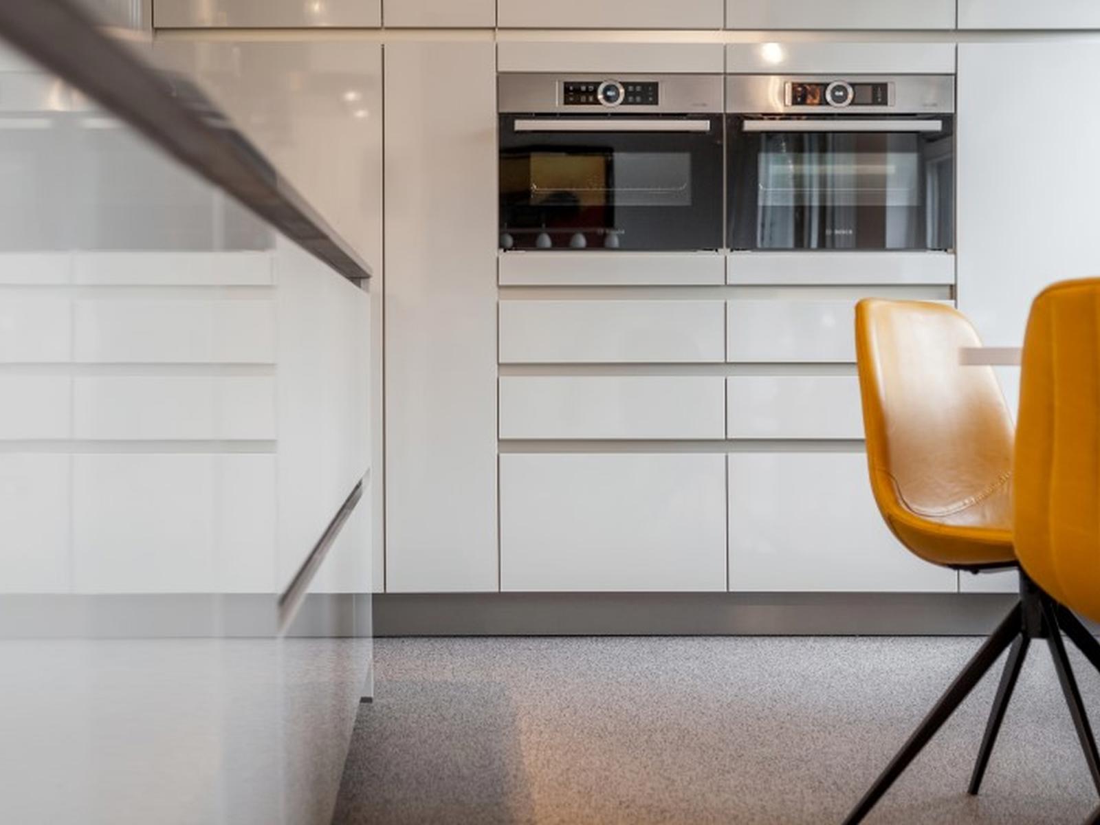 Witte hoogglans greeploze kasten met twee inbouwovens en okergele stoelen