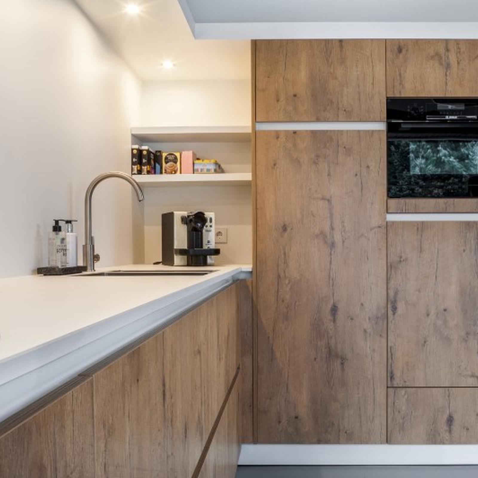 Hoge eikenhouten kastenwand met inbouwoven naast wit werkblad met kraan en koffiemachine