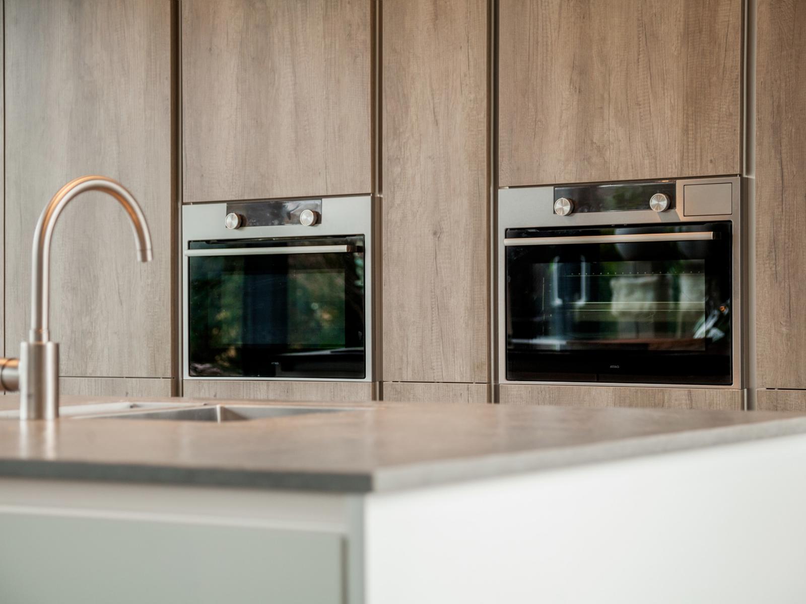RVS kraan op het kookeiland met twee inbouwovens in een houtlook kastenwand op de achtergrond