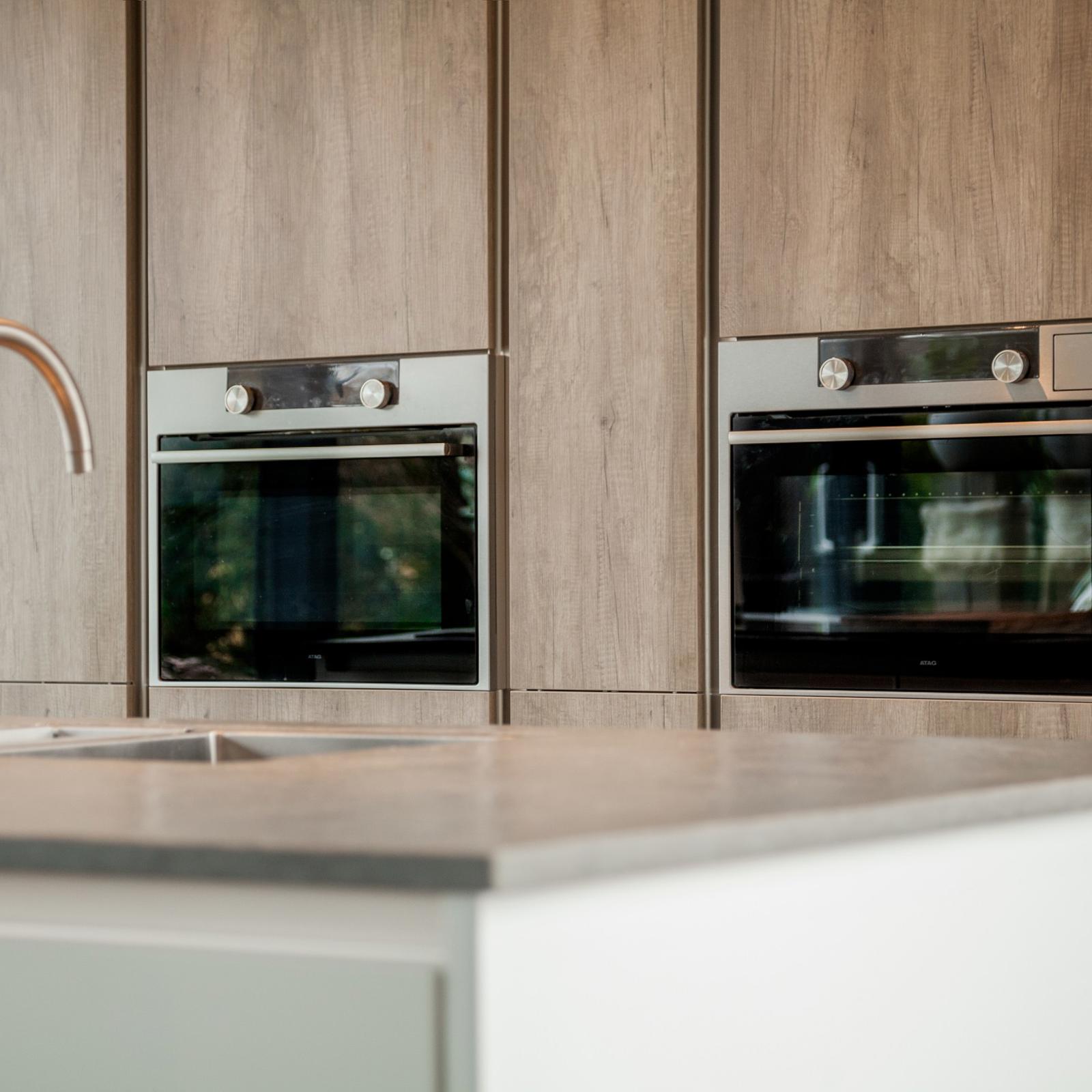RVS kraan op het kookeiland met twee inbouwovens in een houtlook kastenwand op de achtergrond