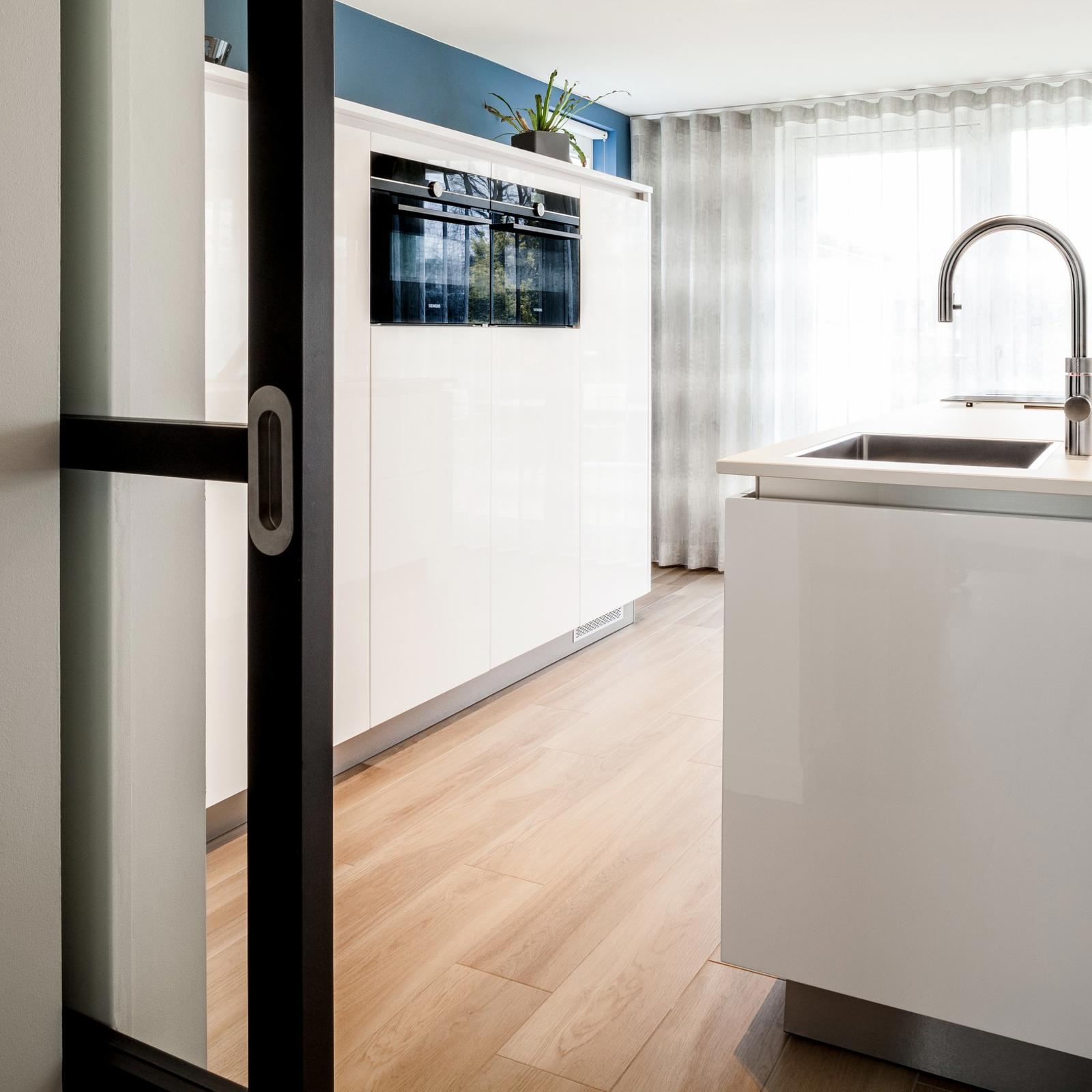 Doorkijk via een zwarte schuifdeur naar het witte hoogglans kookeiland met RVS kraan en een kastenwand met oven