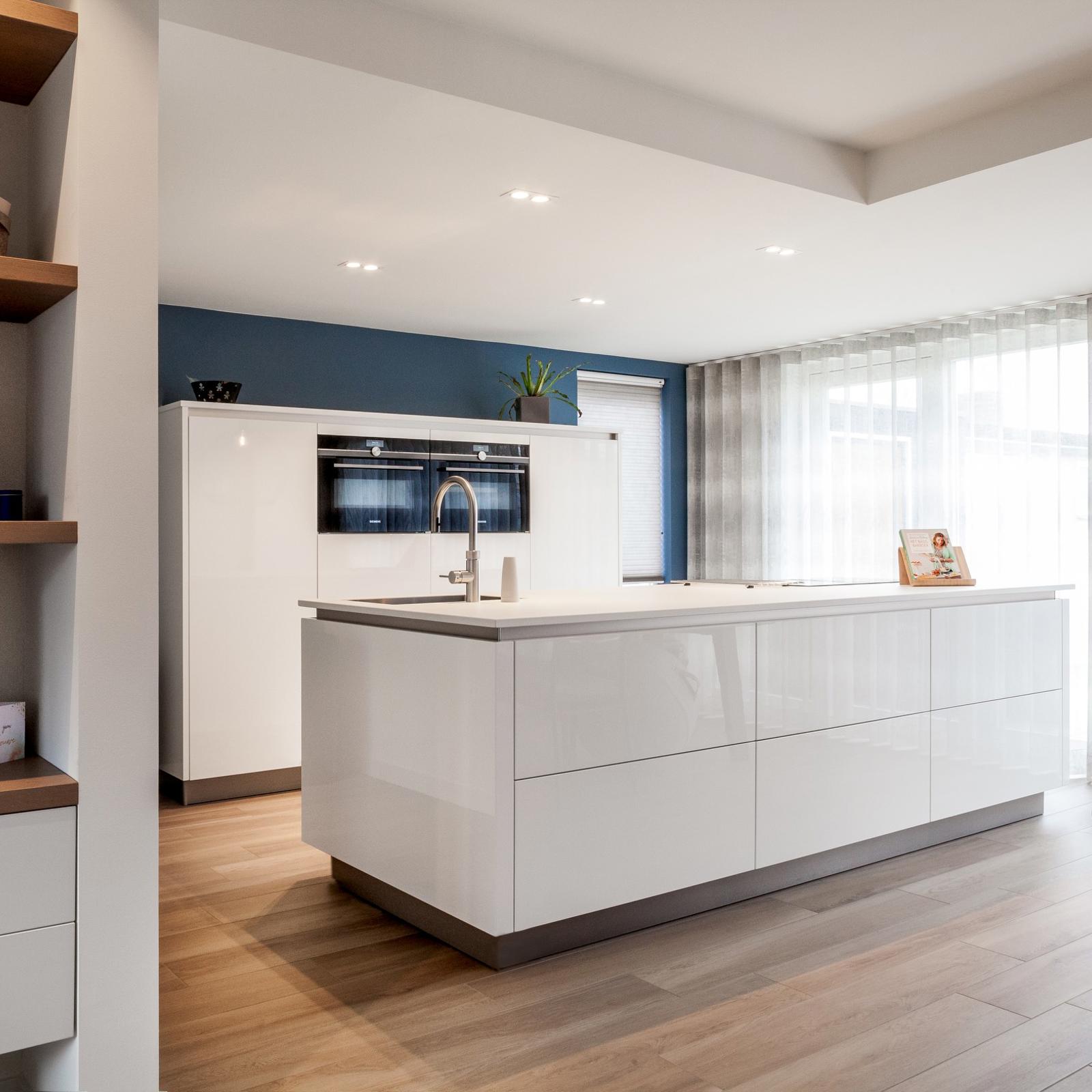 Hoogglans keuken in Beek: wit hoogglans kookeiland voor een kastenwand met inbouwovens en blauwe achterwand