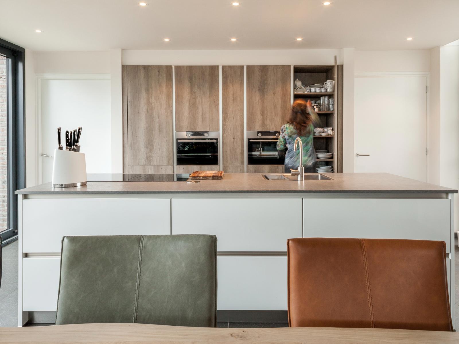 Houtlook keuken in Brunssum: wit kookeiland met eettafel ervoor en een kastenwand in houtlook met open voorraadkast