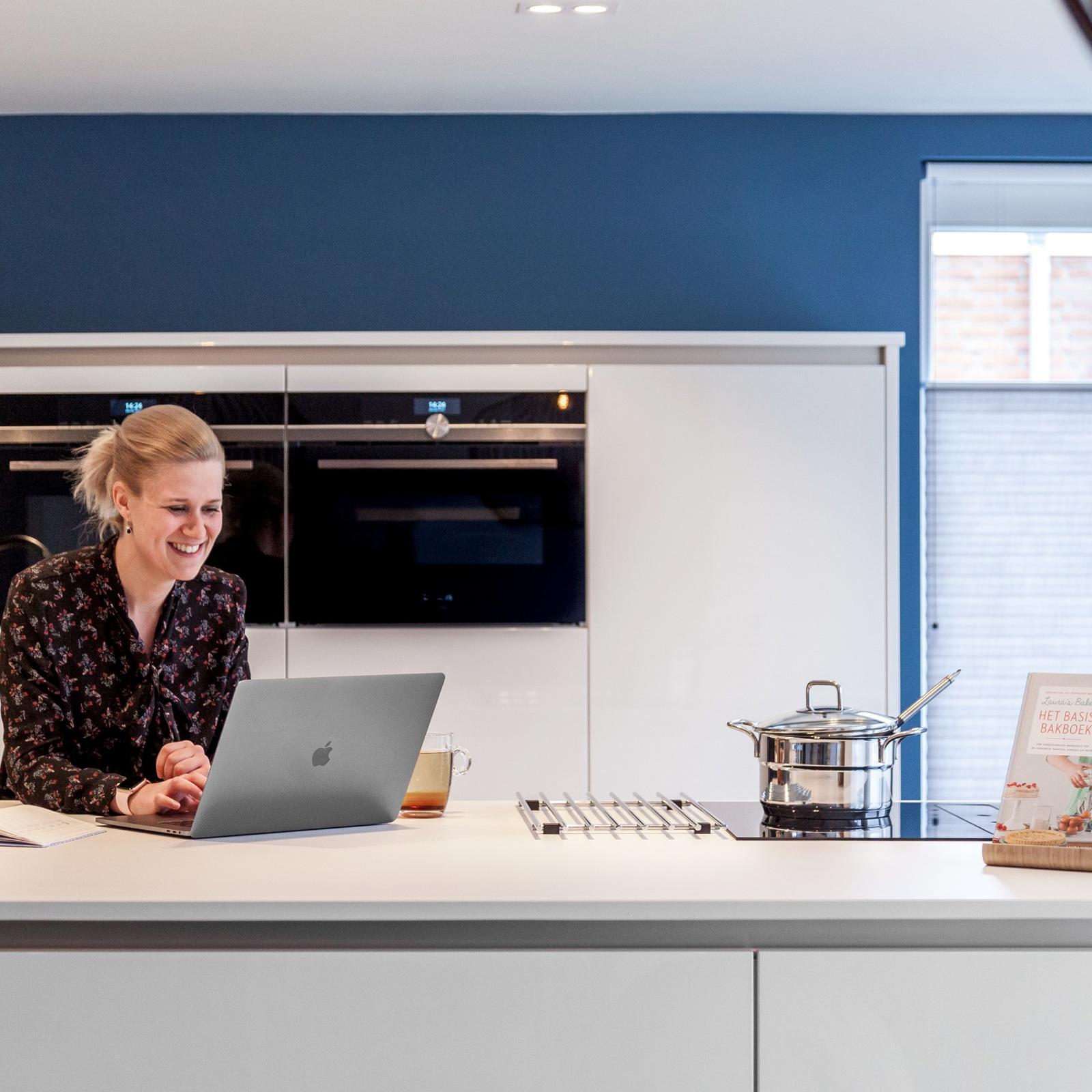 Vrouw met laptop aan een wit kookeiland met kookplaat, voor een witte kastenwand met twee inbouwovens