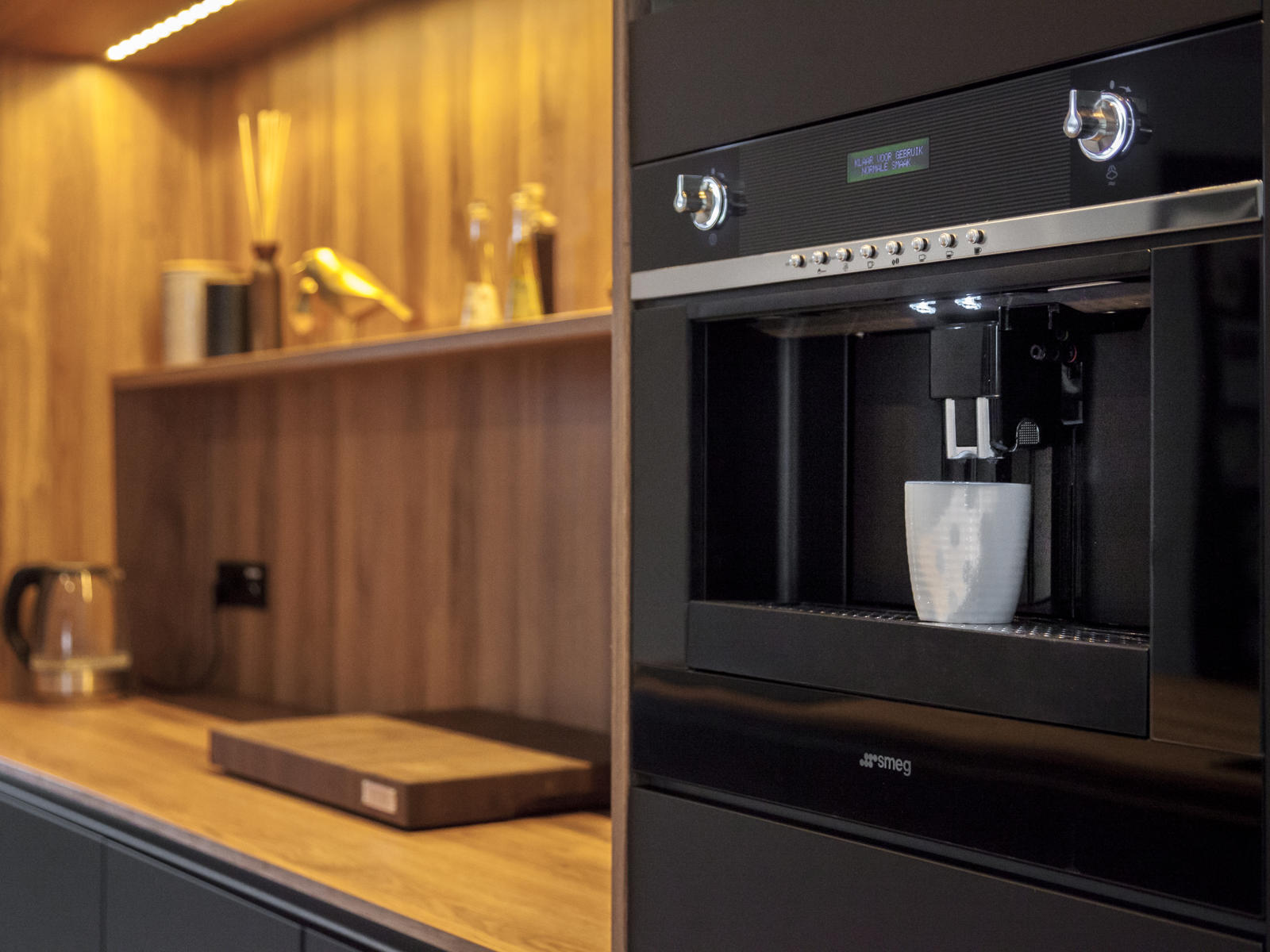 Zwarte Smeg inbouwkoffiemachine naast houten nis met wandplank en werkblad