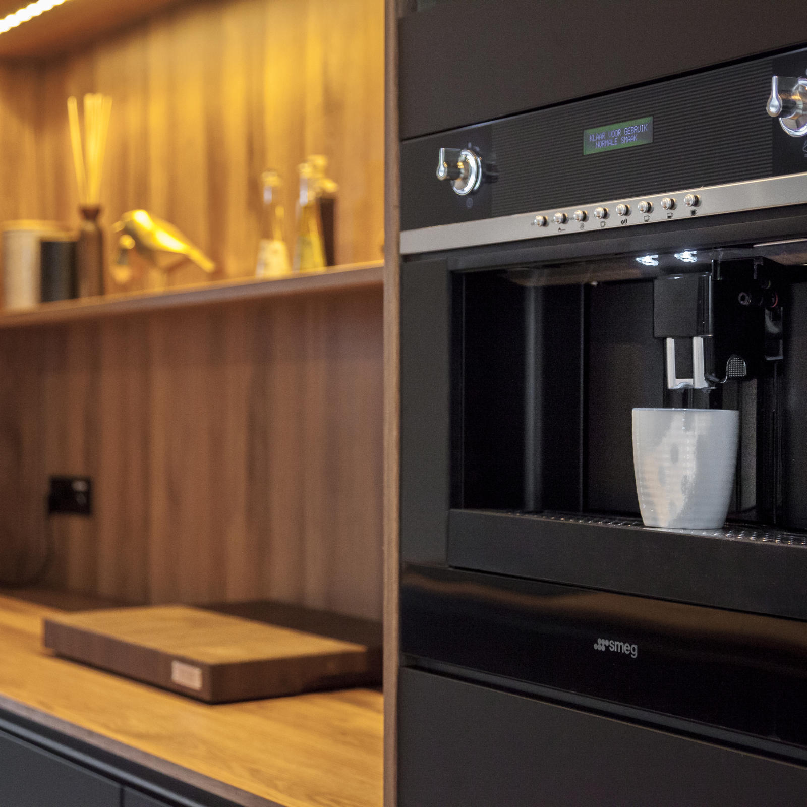 Zwarte Smeg inbouwkoffiemachine naast houten nis met wandplank en werkblad