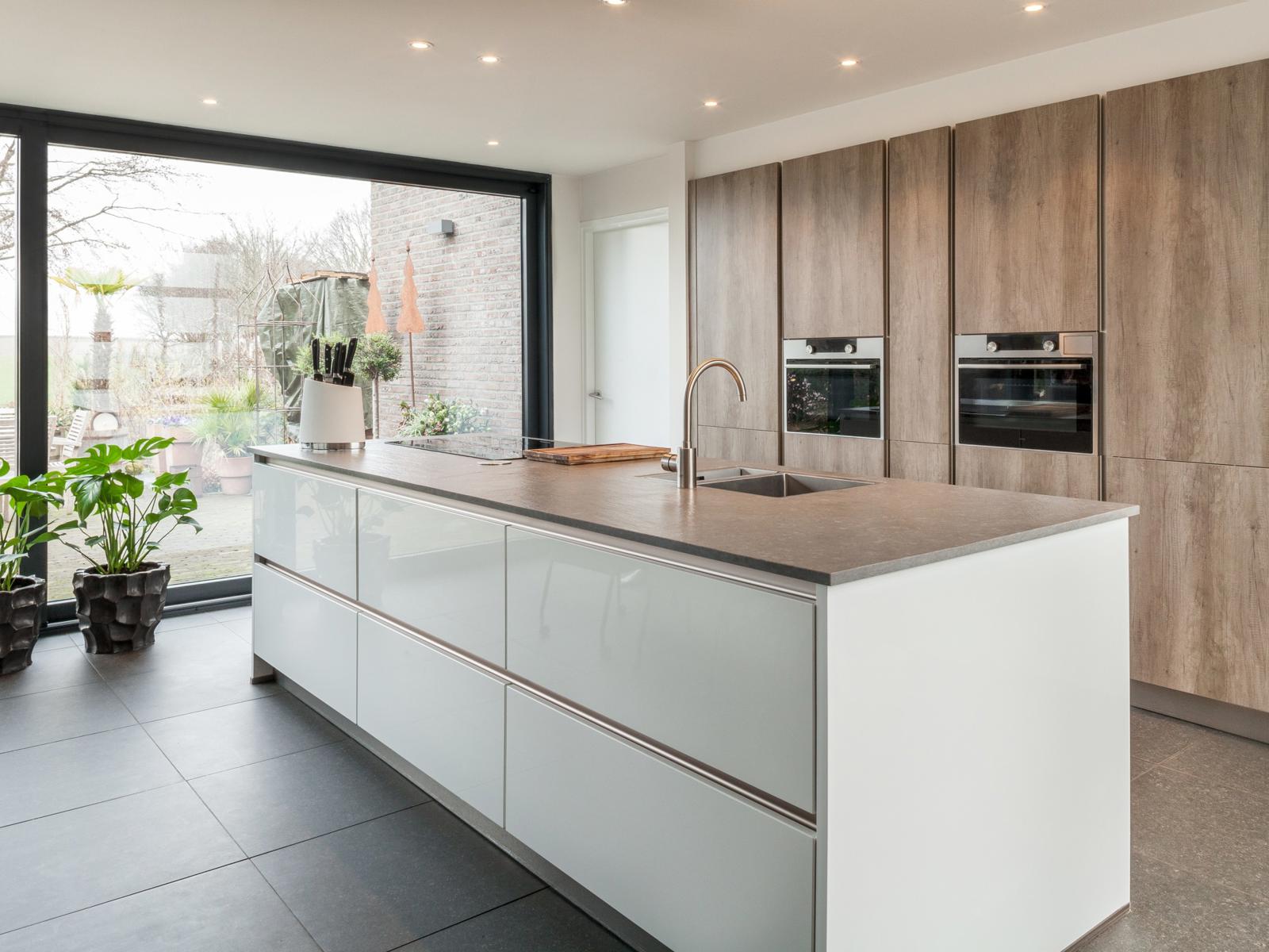 Houtlook keuken in Brunssum: wit hoogglans kookeiland voor een hoge kastenwand in houtlook naast een glazen pui