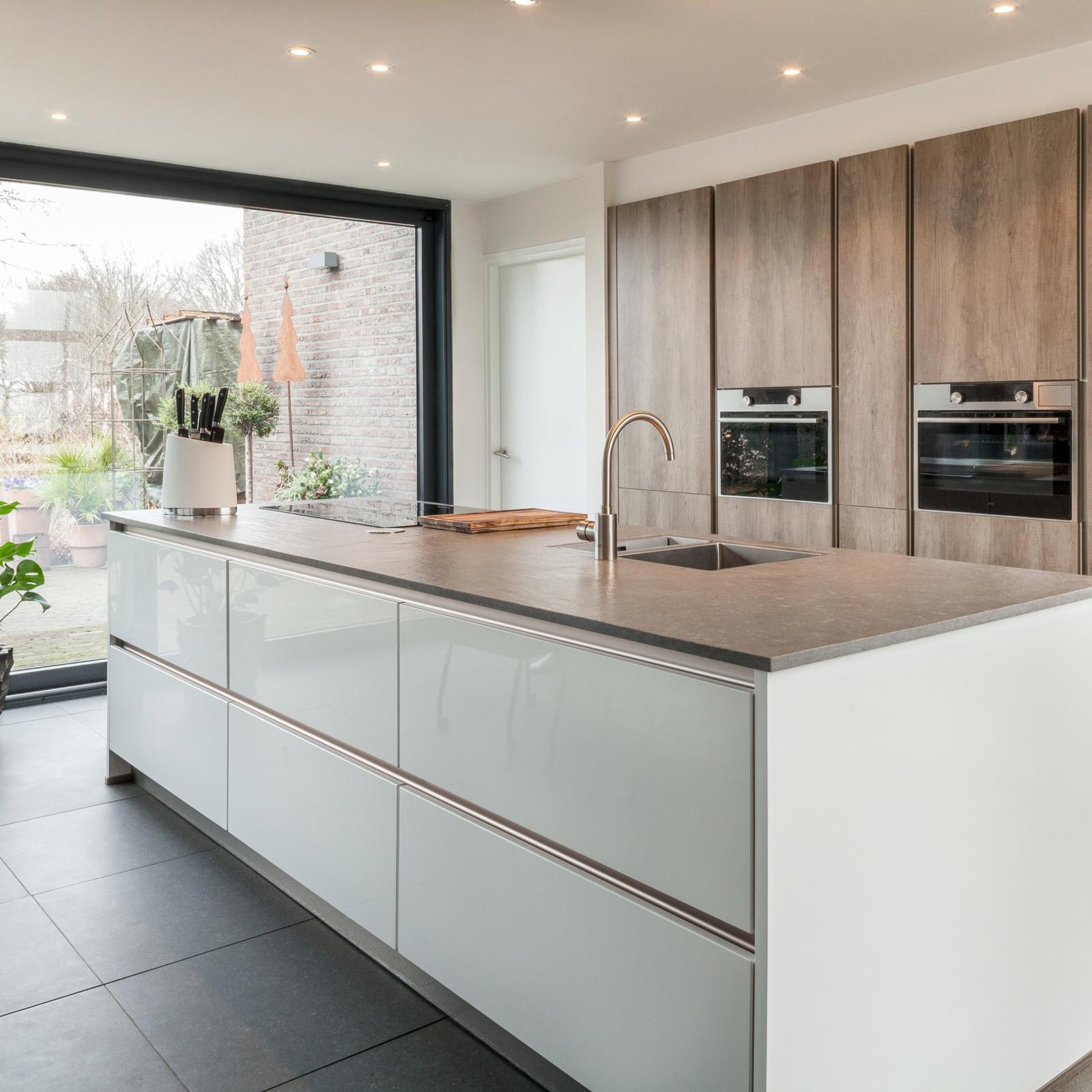 Houtlook keuken in Brunssum: wit hoogglans kookeiland voor een hoge kastenwand in houtlook naast een glazen pui